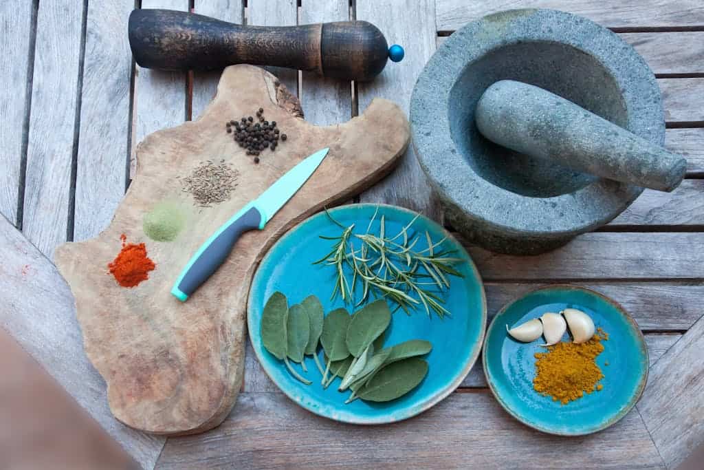 A rustic setup for spice preparation with herbs, spices, garlic, and kitchen tools on wooden table.